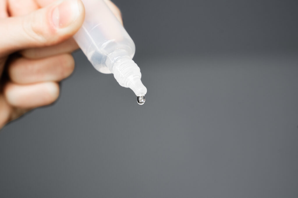 Drop hanging from the transparent tip of a small plastic eye drop or ear drop bottle just before it falls. Hand holding plastic bottle of medicine with droplet. Isolated against a gray background. Important Update Regarding the FDA Eye Drop Recall